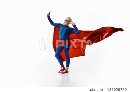 Relaxed, grey-haired man in superhero costume with red cape and sunglasses, making peace sign and taking selfie against white studio background 122056720