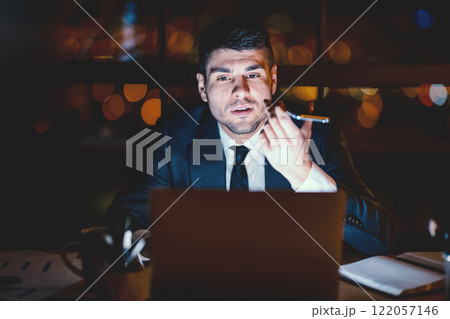 Businessman Using Voice Assistant On Mobile Phone Sitting At Laptop In Modern Office At Night. Selective Focus Businessman Using Voice Assistant On Mobile Phone Sitting At Laptop In Modern Office At Night. Selective Focus 122057146
