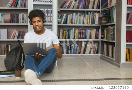 Young african american student with headset sitting on the floor, leaning on bookshelf and using laptop at library, free space Young african american student with headset sitting on the floor, leaning on bookshelf and using laptop at library, free space 122057150