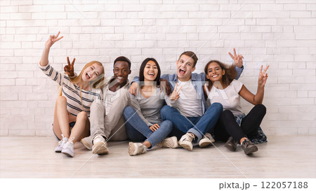 Emotional multiracial students sitting on floor, showing peace gesture, posing at camera, white wall background 122057188
