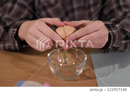 Close-up of hands cracking an egg over a glass bowl, preparing ingredients for baking on a wooden table. Close-up of hands cracking an egg over a glass bowl, preparing ingredients for baking on a wooden table. 122057198