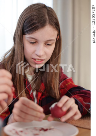 A young girl focused on peeling a red dyed Easter egg over a white plate with scattered eggshells during a festive activity. A young girl focused on peeling a red dyed Easter egg over a white plate with scattered eggshells during a festive activity. 122057207