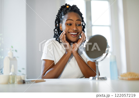 African American young woman with braids is sitting at a table, looking in a mirror with a big smile on her face. She is holding her hands to her cheeks. 122057219