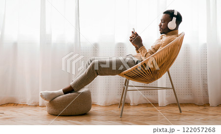 Weekend. Relaxed Black Man In Wireless Headset Using Mobile Phone Listening To Audiobook Sitting On Modern Chair Against Window Indoor Weekend. Relaxed Black Man In Wireless Headset Using Mobile Phone Listening To Audiobook Sitting On Modern Chair Against Window Indoor 122057268