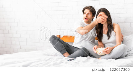 A couple is sitting on a bed watching a movie. The man is holding his arm around the woman, who has her hand over her eyes. Both of them appear scared and shocked. They are eating popcorn 122057300