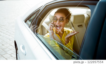 A woman with curly hair and sunglasses is sitting in the back seat of a white car. She is on a phone call and looks excited, with her mouth open in surprise. A woman with curly hair and sunglasses is sitting in the back seat of a white car. She is on a phone call and looks excited, with her mouth open in surprise. 122057554