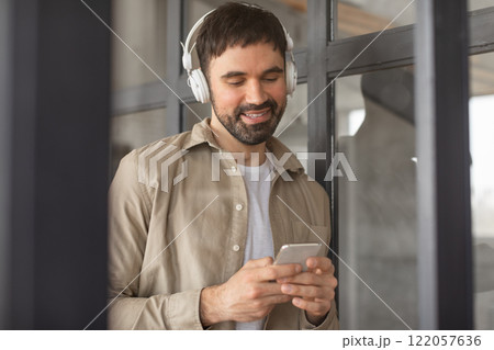 A man in a tan shirt and white t-shirt is standing in an office setting. He is wearing headphones and looking down at his smartphone. He appears to be listening to something on his phone. A man in a tan shirt and white t-shirt is standing in an office setting. He is wearing headphones and looking down at his smartphone. He appears to be listening to something on his phone. 122057636