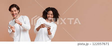 Happy interracial couple playing video games on smartphones, competing each other, standing over yellow studio background Happy interracial couple playing video games on smartphones, competing each other, standing over yellow studio background 122057671
