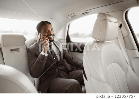 Black businessman in a suit is sitting in the back seat of a car while talking on his phone. He has a serious expression on his face and is looking to the side 122057757