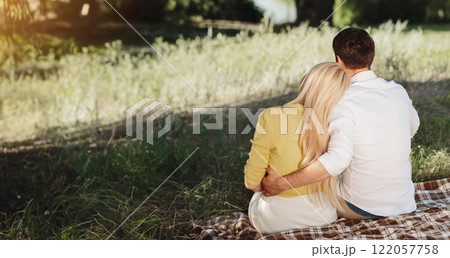 Couple sitting in summer park, having picnic on blanket. Lovers hugging and looking at lake 122057758