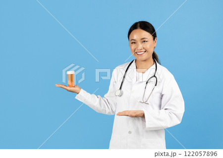 A smiling Asian lady healthcare professional in a white coat presents a prescription bottle while standing against a blue background. A smiling Asian lady healthcare professional in a white coat presents a prescription bottle while standing against a blue background. 122057896