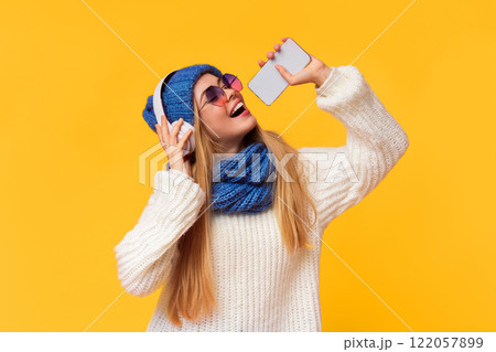 Joyful young winter girl in wireless headphones listening to music and singing in phone over pink background Joyful young winter girl in wireless headphones listening to music and singing in phone over pink background 122057899