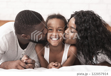 Portrait of loving african american parents kissing cheeks of their happy little daughter, closeup 122057914