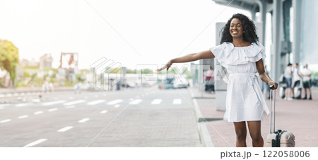 A young African American woman in a white dress smiles happily while hailing a taxi outside an airport. She is pulling a suitcase behind her. The airport is in the background, copy space A young African American woman in a white dress smiles happily while hailing a taxi outside an airport. She is pulling a suitcase behind her. The airport is in the background, copy space 122058006