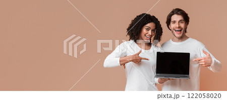 Cool website. Smiling multiracial couple holding laptop with black screen and pointing at it, posing together over yellow background with empty space 122058020