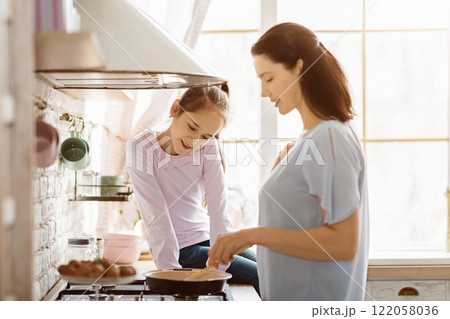 A young girl is learning to cook with her mother in a modern kitchen. The girl is standing at the stove, stirring something in a pan, while her mother watches and smiles A young girl is learning to cook with her mother in a modern kitchen. The girl is standing at the stove, stirring something in a pan, while her mother watches and smiles 122058036