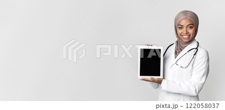 Online pharmacy concept. Black muslim female doctor holding digital tablet with black blank screen for mockup, posing over gray background, free space 122058037
