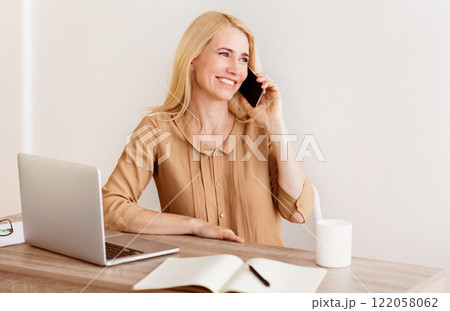 A woman with blonde hair is sitting at a desk, talking on her phone and smiling. A laptop and a notebook are on the desk in front of her. A woman with blonde hair is sitting at a desk, talking on her phone and smiling. A laptop and a notebook are on the desk in front of her. 122058062