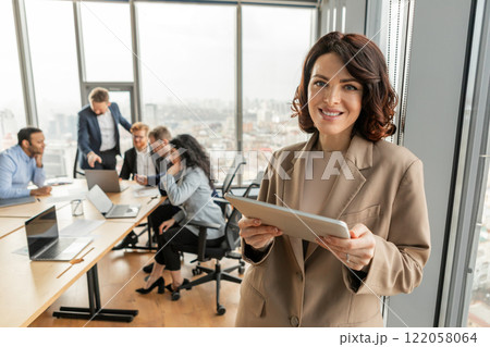 A businesswoman in a beige blazer stands in a modern office, holding a tablet and smiling at the camera. She is positioned near a large window with a view of a cityscape. A businesswoman in a beige blazer stands in a modern office, holding a tablet and smiling at the camera. She is positioned near a large window with a view of a cityscape. 122058064