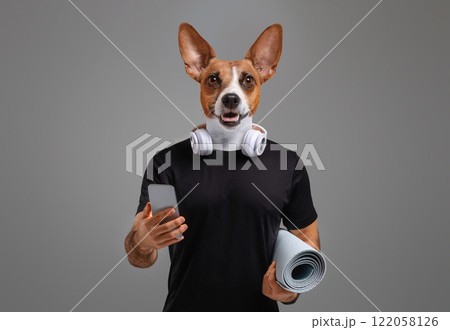 A person with a dog's head is wearing a black t-shirt and holding a rolled-up yoga mat in one hand and a smartphone in the other. They are wearing headphones around their neck. 122058126