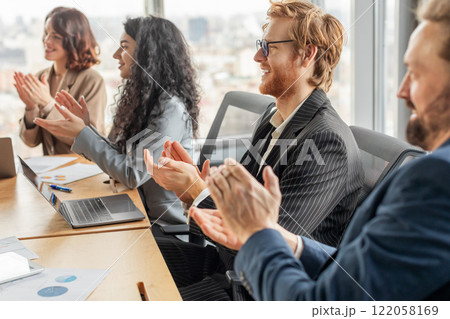 A group of business professionals sit at a table in a modern office, clapping their hands in appreciation as they listen to a presentation. They are dressed in formal attire 122058169