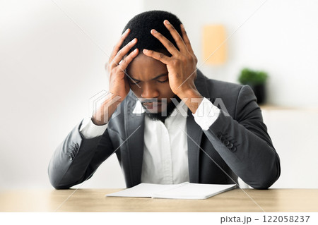 A businessman in a suit sits at a desk with his head in his hands, looking down at paperwork. 122058237