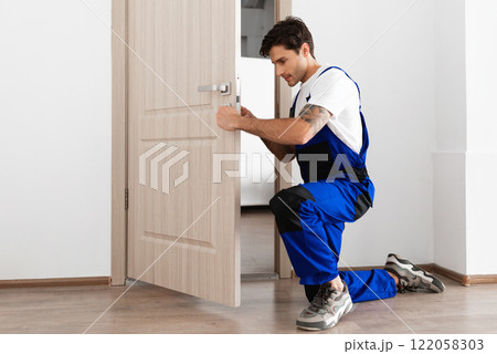 Installation of a lock on the front wooden entrance door. Portrait of young locksmith workman in blue uniform installing door knob. Professional repair service. Maintenance Concept Installation of a lock on the front wooden entrance door. Portrait of young locksmith workman in blue uniform installing door knob. Professional repair service. Maintenance Concept 122058303