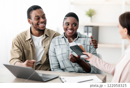 Travel Agent Giving Tickets To Happy Black Travelers Couple Sitting In Tour Agency Office. Tourism Management And Insurance Service, Family Vacation Tour Concept. Selective Focus 122058325