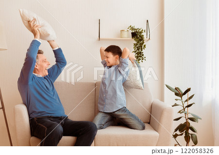 Sleepover With Grandparent. Happy Boy And His Grandfather Fighting With Pillows Having Fun Together Sitting On Couch At Home 122058326