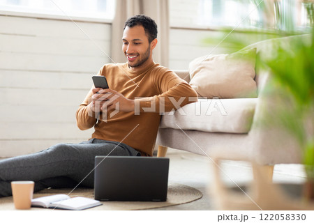 Business And Telecommuting Concept. Portrait of smiling young Arabic male using smartphone sitting on the floor with laptop computer, reading text message, leaning on sofa, working at home office Business And Telecommuting Concept. Portrait of smiling young Arabic male using smartphone sitting on the floor with laptop computer, reading text message, leaning on sofa, working at home office 122058330