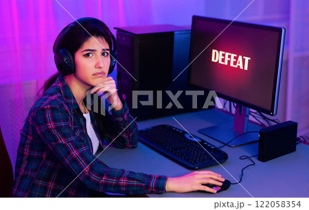 A young woman, wearing headphones, sits at a computer desk with a serious expression on her face. The word DEFEAT is displayed on the computer screen, indicating that she has lost a game A young woman, wearing headphones, sits at a computer desk with a serious expression on her face. The word DEFEAT is displayed on the computer screen, indicating that she has lost a game 122058354