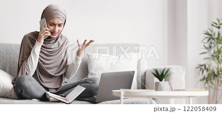 Freelance stress. Concerned muslim girl in hijab talking on cellphone and emotionally gesturing while working online on laptop at home, free space Freelance stress. Concerned muslim girl in hijab talking on cellphone and emotionally gesturing while working online on laptop at home, free space 122058409