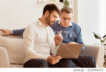 Computer Literacy. Adult Son Teaching Aging Dad How To Use Laptop And Internet Sitting On Couch Indoor Computer Literacy. Adult Son Teaching Aging Dad How To Use Laptop And Internet Sitting On Couch Indoor 122058413