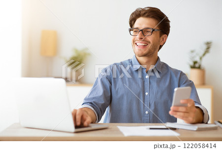 A man wearing glasses sits at a desk working on his laptop, smiling as he holds a phone in his other hand. 122058414