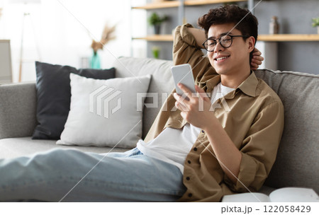 Enjoying Weekend. Handsome Asian Man In Glasses Using Smartphone While Sitting Leaning On Couch At Home, Happy Guy Relaxing In Living Room, Browsing Internet Or Messaging With Friends, Selective Focus 122058429