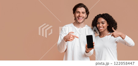 Cool app. Smiling interracial couple holding and pointing at modern smartphone with black screen for mockup, posing together on yellow background Cool app. Smiling interracial couple holding and pointing at modern smartphone with black screen for mockup, posing together on yellow background 122058447