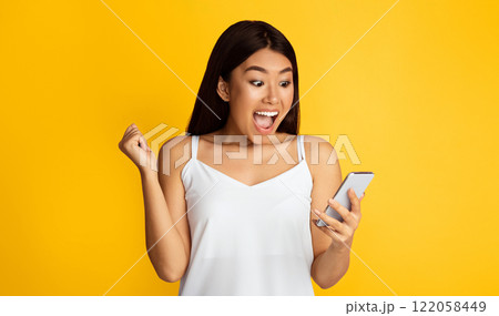 A young Asian woman, wearing a white tank top, is standing in front of a bright yellow background. She is holding a smartphone in her right hand, looking at it with excitement 122058449