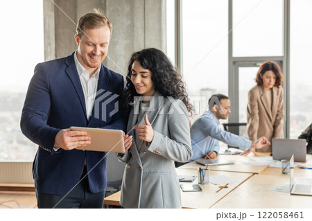 Two business professionals are working together in a modern office setting. They are standing near a large wooden table with other colleagues in the background. The man is holding a tablet 122058461