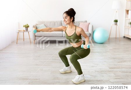 Happy young Indian woman doing squats with dumbbells at home, full length. Positive millennial Asian lady performing strength exercises, keeping in good shape during covid quarantine 122058574