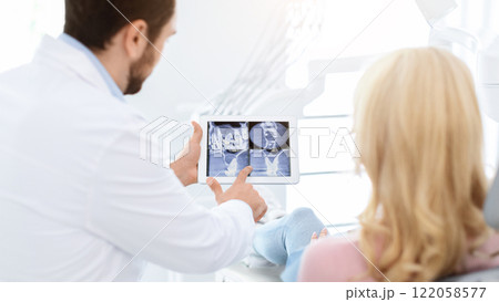 Man dentist showing female patient treatment goal, using digital tablet, dental clinic interior. Bearded man stomatologist having conversation with lady in dental chair, pointing at gadget, back view 122058577