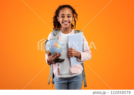 Portrait of cheerful young black girl holding Earth world globe and notebook, wearing backpack and looking at camera isolated on yellow color background at studio. Education in school concept 122058579