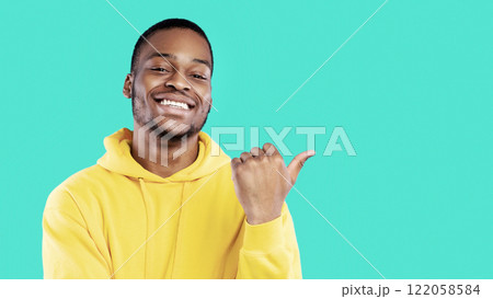 Smiling African Guy Pointing Thumb Finger Aside At Blank Space For Text Advertising Your Offer Standing Over Blue Background, Studio Shot. Look There Concept. Panorama 122058584