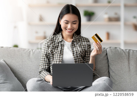 Cheerful young asian woman sitting on couch in living room and using laptop and credit card, checking new webstore for online shopping or banking, home interior, copy space, catching sales Cheerful young asian woman sitting on couch in living room and using laptop and credit card, checking new webstore for online shopping or banking, home interior, copy space, catching sales 122058659