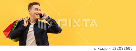 Positive handsome young guy wearing casual clothing with colorful shopping bags on his shoulder talking on phone and looking at copy space for advertisement, yellow background, web-banner 122058693