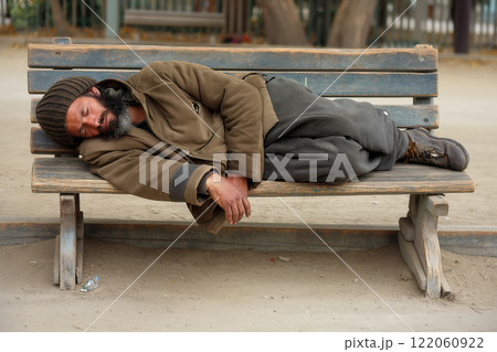 Poor homeless man or refugee sleeping on the wooden bench on the urban street in the city 122060922