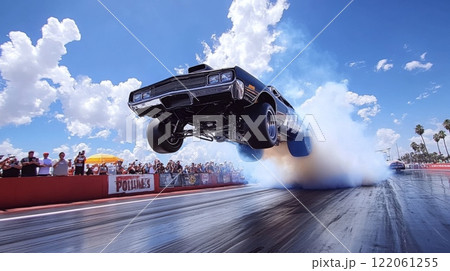 A classic car performs an impressive jump during a drag racing event, surrounded by an enthusiastic crowd and a vibrant blue sky. 122061255