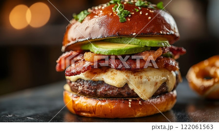 A delicious bacon cheeseburger features a pretzel bun, melted cheese, crispy bacon, fresh avocado, and herbs, served in a warm, inviting restaurant atmosphere. 122061563
