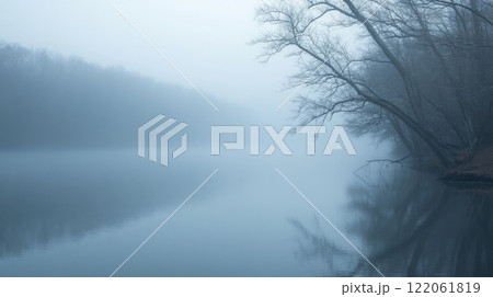 In the early morning, a river is shrouded in thick mist, with reflective calm waters capturing the gray skies. Bare trees line the riverbank, creating a peaceful, almost ethereal atmosphere. 122061819