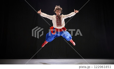 Dancer in embroidered shirt and blue Ukrainian folk attire, jumping wide legs and hands against black background, emphasizing athleticism of Ukrainian Hopak. 122061851