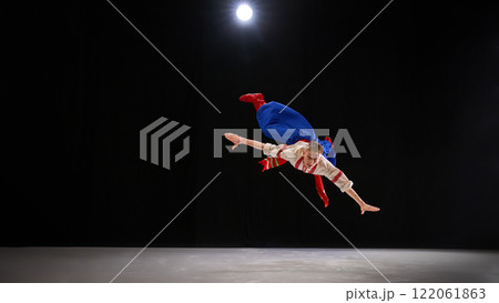 Performer dynamically leaping with arms and legs extended, illustrating energetic Hopak dance movement under focused stage spotlight on black backdrop. 122061863